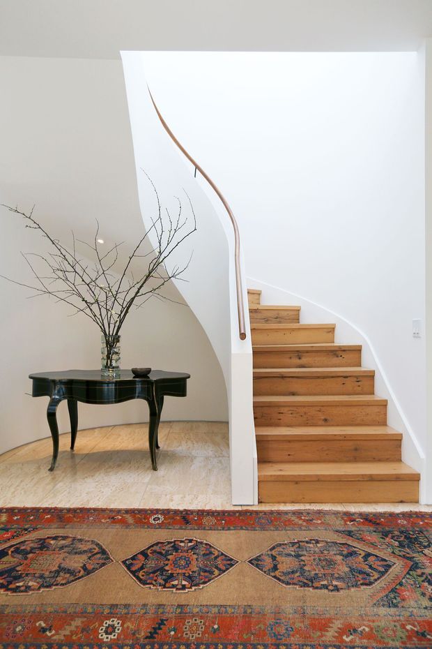 Bright interior view of a sculptural curved staircase with warm timber treads, white plaster walls, and a slim timber handrail. A dark traditional console table with a glass vase and bare branches sits beneath the stair, while layered patterned Persian-style rugs cover the pale floor.