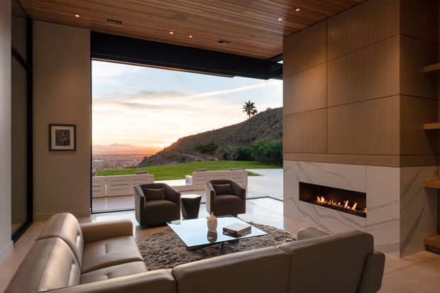 A refined contemporary living room with a large open glazed wall framing a sunset hillside view. The room features a warm timber ceiling, dark structural framing, a large sectional sofa and lounge chairs arranged around a glass coffee table, and a linear built-in fireplace set into a veined pale stone feature wall beneath timber-panelled joinery.