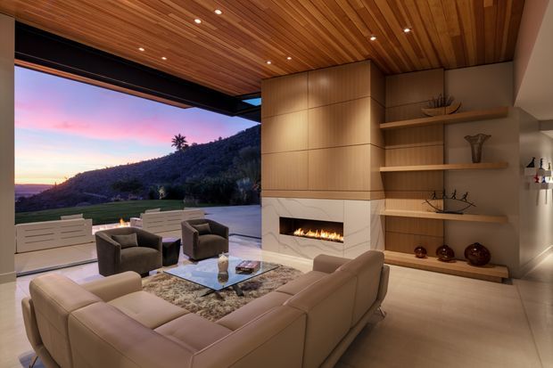 A refined contemporary living room opening onto an expansive outdoor terrace at sunset. The room features a large cream sectional sofa, armchairs, glass coffee table, built-in linear fireplace with a pale stone surround, extensive warm timber wall cabinetry and open shelving, and a slatted timber ceiling with recessed lighting. Full-height open glazing frames a landscaped hillside and sunset beyond.