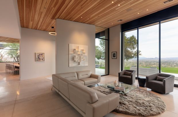 Spacious contemporary living room with a large cream sectional sofa, sculptural coffee table, armchairs, and a textured area rug. The room features expansive floor-to-ceiling glazing overlooking a green landscape, a warm timber-lined ceiling with recessed lighting, pale smooth walls, and polished light flooring. An open dining area is visible beyond the living space.