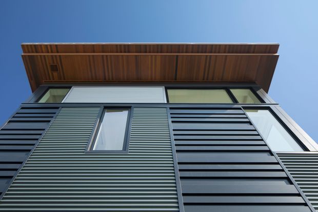 Low-angle close-up of a contemporary building facade against a clear blue sky. The exterior combines dark horizontally ribbed metal cladding, large rectangular windows, and a projecting timber-lined soffit or roof canopy with visible exposed wood grain.