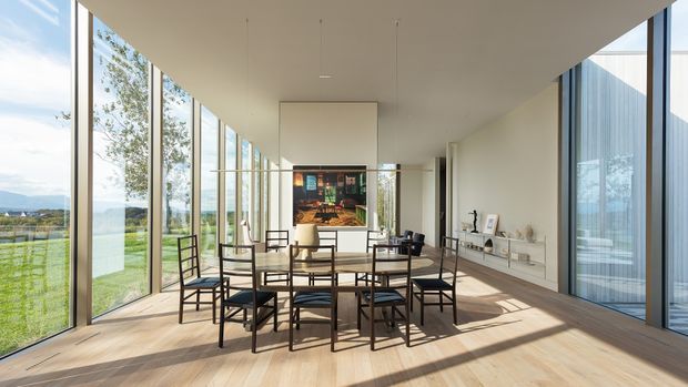 A bright, elongated contemporary dining room with floor-to-ceiling glazing along both sides, a large rectangular dining table surrounded by black-framed chairs, and a minimalist open display shelf along the right wall. Warm timber flooring, white walls and ceiling, slim dark window frames, and linear pendant lighting create a clean, airy interior. The glazing provides views toward a rural or landscaped setting, while a colourful artwork anchors the far wall.