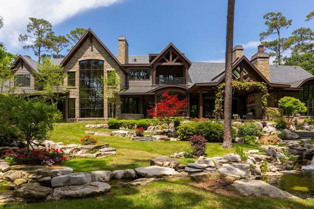 A large multi-wing lodge-style residence set within a landscaped garden, featuring steep gabled roofs, stone-clad walls, extensive dark timber framing, tall glazed openings, multiple masonry chimneys, and covered outdoor connections. The foreground includes layered natural stone edging, planted beds, lawn, and a small pond or watercourse. Bright daylight and mature trees surround the property.