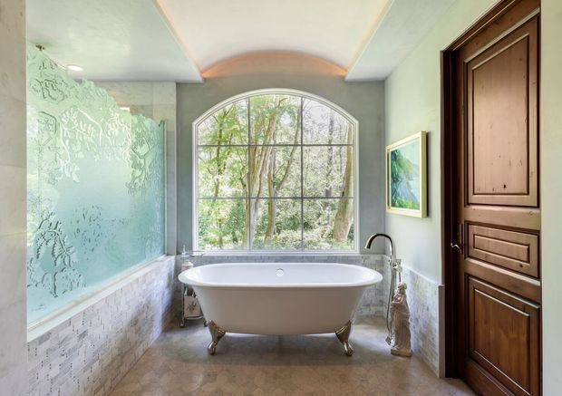 Elegant traditional bathroom featuring a freestanding clawfoot bathtub beneath a large arched divided-light window. The room has pale green walls, a decorative patterned glass shower enclosure on the left, light mosaic wall tile, a stone-look floor, a sculpted ceiling detail, and a dark stained timber paneled door. A vintage-style bathtub and traditional fittings create a refined, spa-like atmosphere.