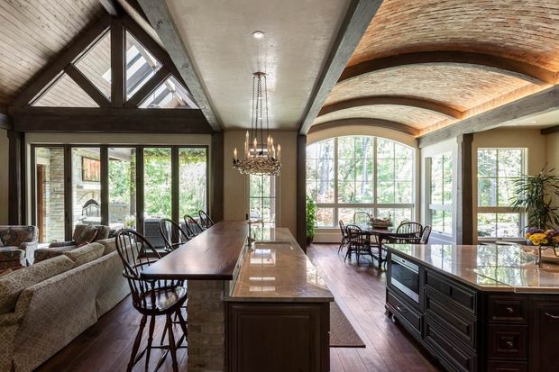 A spacious open-plan kitchen and dining area with a large dark timber island, bar seating, and extensive glazing. The room combines exposed dark structural beams, warm brick barrel-vault ceiling sections, brick walls, timber flooring, and traditional multi-pane windows. A living area with a patterned sectional sofa is visible to the left, while a dining table occupies the glazed area beyond the kitchen.
