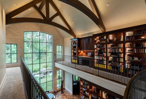 A grand double-height library or study viewed from an upper-level landing, featuring extensive dark timber built-in bookcases, a mezzanine-style gallery with black metal balustrades, tall arched multi-pane windows, exposed dark timber trusses, pale brick walls, and a parquet floor visible below. The room is warmly lit and has a refined traditional character.
