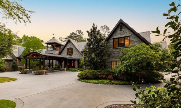 Exterior view of a large, multi-building residence with steep gabled roofs, dark grey stone cladding, timber-framed windows, and a covered porte-cochère over the driveway. Mature landscaped gardens surround the broad paved approach, with a prominent central gable and smaller connected wings.