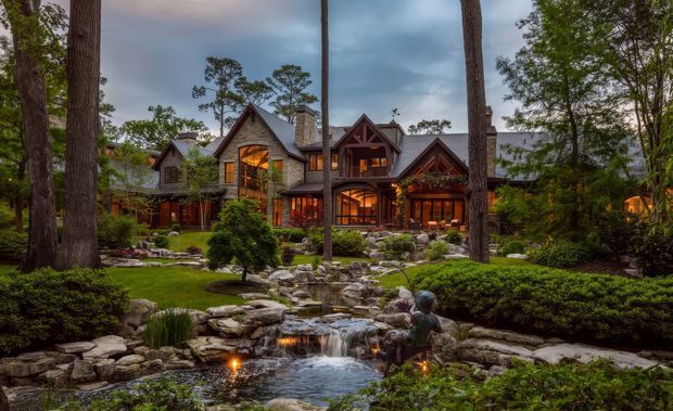 A large rustic lodge-style house nestled among mature trees, photographed at dusk. The facade features multiple steep gables, dark timber framing, stone-clad walls and chimneys, with warm light glowing through the windows. A landscaped garden with stone edging, shrubs, a pond and a small cascading stream fills the foreground.