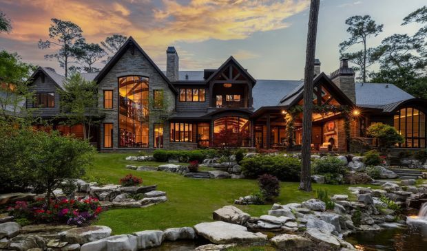 Exterior view of a large lodge-style residence set within a landscaped garden at dusk. The home features multiple steep gables, dark grey shingle-like cladding, extensive warm-lit glazing, timber framing, prominent stone walls and chimneys. A landscaped water feature with rocks and a small waterfall occupies the foreground, with mature trees and garden planting surrounding the building.