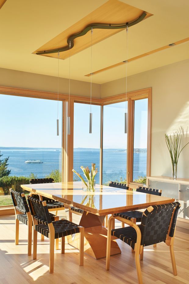 Bright dining room with a large polished timber dining table and woven black-and-timber dining chairs, positioned beside expansive corner windows overlooking a blue waterfront. Timber-framed glazing, pale timber flooring, cream walls, and a sculptural timber ceiling feature with suspended pendant lights create a warm, airy interior.