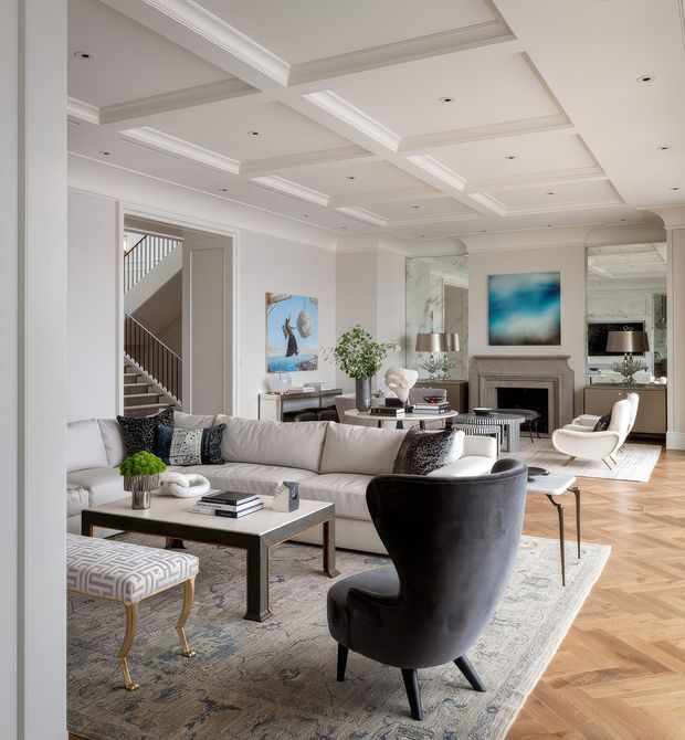 A bright, refined living room with a large cream sectional sofa, black accent armchair, patterned ottoman, coffee table, and neutral area rug. The room features a decorative fireplace with mirror panels, contemporary artwork, herringbone timber flooring, and a highly detailed coffered ceiling with recessed lighting. A staircase is visible through an open doorway.