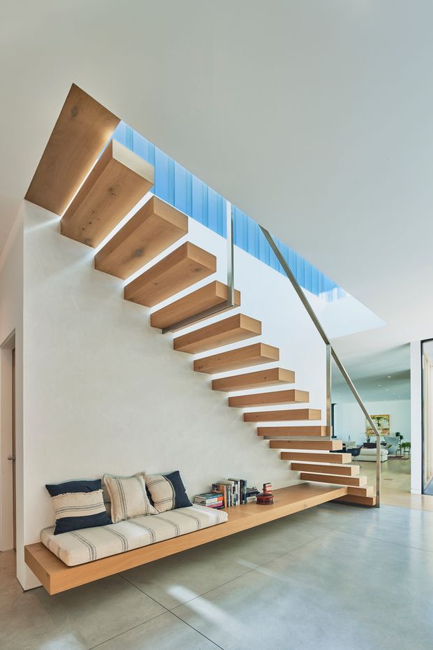 Bright contemporary interior featuring a floating open-riser staircase with thick timber treads, slim metal balustrade, and a large integrated timber bench with upholstered seating beneath. The staircase rises beside a white plaster wall toward a skylit or clerestory-lit void, with a polished grey concrete floor and a partially visible open-plan living area beyond.