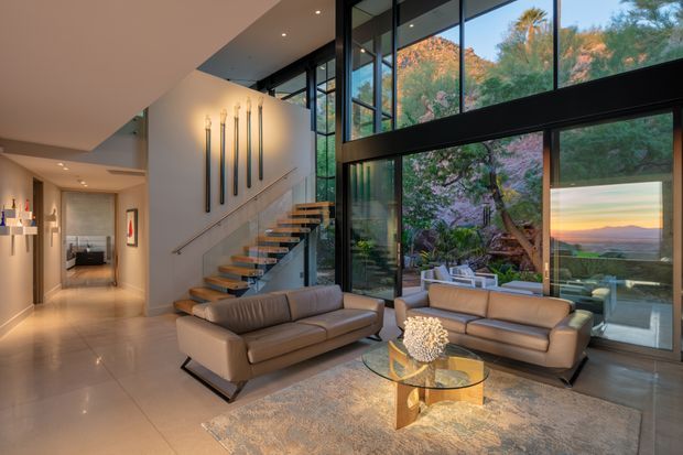 A sophisticated contemporary living room with a high glazed wall overlooking a landscaped garden and distant sunset landscape. The space features a floating timber-and-metal staircase, polished light flooring, neutral sofas, a round glass coffee table, recessed lighting, and tall sculptural wall lights. Floor-to-ceiling black-framed glazing creates a strong indoor-outdoor connection and abundant natural light.