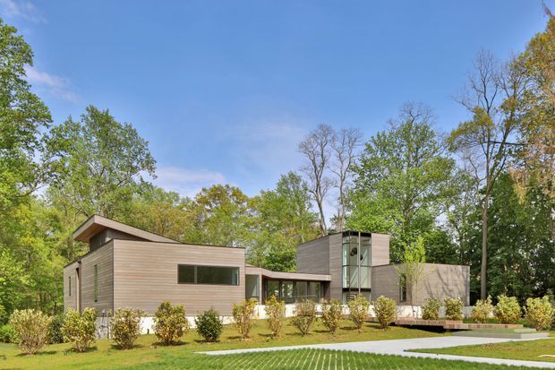 Exterior view of a contemporary, low-profile residential house set within a landscaped, tree-filled site. The home has horizontal timber-toned cladding, simple rectilinear volumes, broad glazing, and a prominent glazed vertical stair or circulation element. A paved pathway and manicured planting lead toward the entrance.