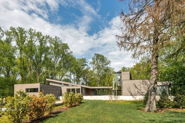 A low, modern single-storey residence set within a landscaped, tree-filled site. The building has a flat roofline, horizontal massing, extensive glazing, and a restrained palette of dark timber-toned and light neutral wall surfaces. A broad lawn, clipped hedges, mature deciduous trees, and a partly enclosed courtyard-like arrangement surround the house under a bright sky.