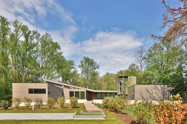 Low-profile modern single-storey residence set within a wooded suburban landscape. The home has horizontal timber-toned cladding, broad rectangular windows, a long low roofline, and a taller central volume, with a stepped concrete path leading through landscaped planting to the entrance.