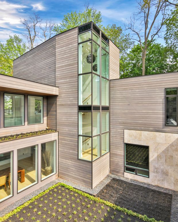 Exterior view of a contemporary multi-wing home arranged around a landscaped courtyard. The building has horizontal wood-toned cladding, extensive black-framed floor-to-ceiling glazing forming a tall central vertical feature, and a light natural-stone wall section. The courtyard contains newly planted garden beds and gravel paths, with mature trees surrounding the house.