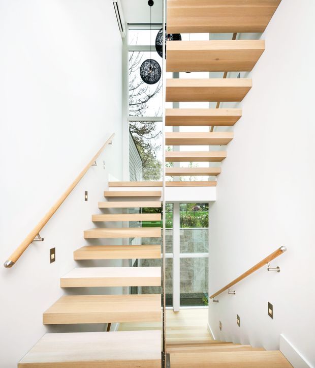 Bright modern stairway with floating light-wood treads ascending through a tall glazed void. White walls, slim glass balustrade, timber handrails, and large windows create an airy vertical circulation space, with black spherical pendant lights and greenery visible outside.