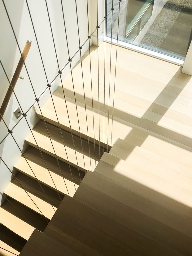 A bright modern stairway viewed from above, with pale timber treads and stringers, a white wall, and slender dark vertical cable balustrade. Strong sunlight enters through large glazing beside the stairs, casting geometric shadows across the steps and landing.