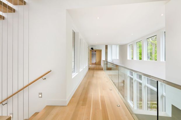 Bright modern upper-level hallway or landing with pale timber flooring, white walls and ceiling, recessed downlights, and extensive floor-to-ceiling glazing. A glass balustrade overlooks a lower level, while a simple timber handrail lines the stair edge at left. The space has a long, open linear plan and abundant natural light.