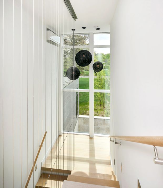 Bright contemporary stairway with pale timber treads, white walls, a timber handrail, and a distinctive vertical cable balustrade. Large floor-to-ceiling windows bring in abundant natural light and overlook a landscaped garden. Three dark spherical pendant lights hang over the stair landing, creating a strong visual contrast.