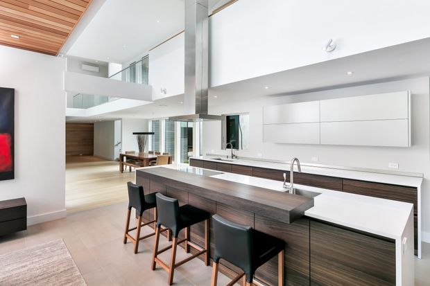 A bright, open-plan contemporary kitchen viewed from the island, with dark timber cabinetry, a long stone island and integrated white benchtop extension seating four stools. The kitchen opens to a double-height living area with a mezzanine, extensive glazing, white walls, and a warm timber slatted ceiling. A large wall-mounted white display or storage unit and linear lighting contribute to the clean modern aesthetic.