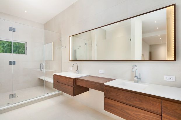 Bright contemporary bathroom with a large frameless glass walk-in shower, double vanity, wall-mounted dark timber cabinetry, white benchtops, and a wide framed mirrored cabinet. Pale neutral wall and floor finishes create a clean, spacious appearance.