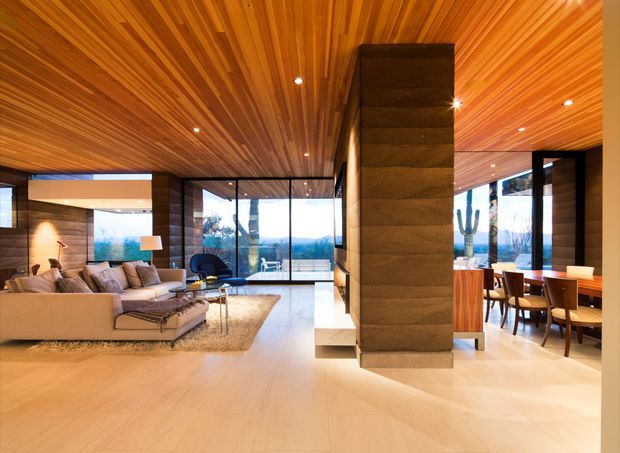 A spacious open-plan contemporary interior combining a living room and dining area. The room features extensive warm timber slat lining across the ceiling and feature walls, pale smooth flooring, a large sectional sofa, a central dark stone-clad structural column, full-height black-framed glazing, and a timber dining table. The interior is bright, warm, refined, and strongly connected to the outdoor landscape visible through the glazing.
