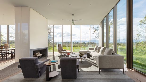 A bright, spacious contemporary living room with floor-to-ceiling glazing on multiple sides, overlooking a landscaped rural or suburban setting. The room features a large neutral sectional sofa, dark lounge chairs, a low coffee table, a fireplace set into a pale wall, timber flooring, a ceiling fan, and understated recessed lighting. An adjoining dining area is visible at left.
