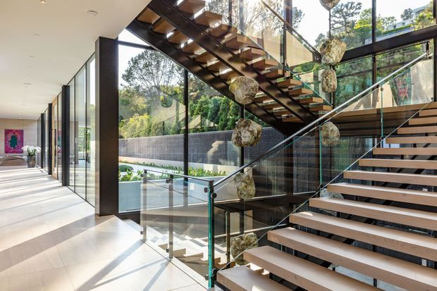 Bright contemporary interior centered on a sculptural open-riser staircase with dark timber treads, slim dark structural elements, and clear glass balustrades. Large floor-to-ceiling windows line the circulation space, bringing in abundant daylight and views of landscaped grounds. Decorative suspended stone-like forms hang beside the stair, while a light tiled floor and white ceiling create a clean, airy gallery-like setting.