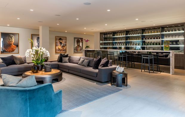 A sophisticated open-plan living and home bar area. A large grey sectional sofa and bright blue lounge chair surround a round coffee table, with artwork and orchids adding decorative detail. The rear of the room features a substantial backlit bar with extensive bottle display shelving, a stone or light-coloured counter, and high stools. Recessed ceiling lighting and pale flooring create a polished, inviting atmosphere.
