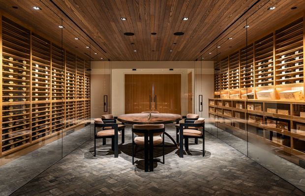 A sophisticated wine room with extensive floor-to-ceiling wine storage lining both side walls, illuminated display shelving, a central round timber table with upholstered dining chairs, glass partitions, a warm timber-lined ceiling, and dark stone or concrete-look flooring. The room has a luxurious, intimate, gallery-like atmosphere with carefully integrated recessed and pendant lighting.
