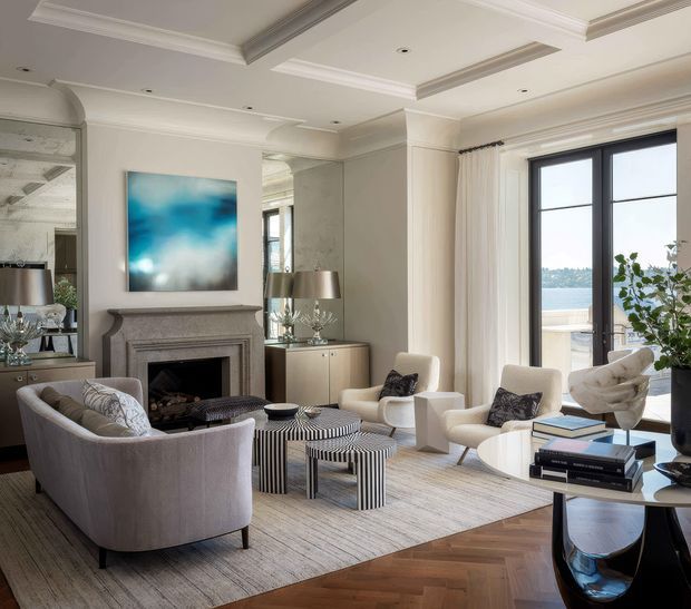 Elegant, light-filled formal living room with a sculpted grey fireplace, large abstract artwork, mirrored wall panels, a pair of cream lounge chairs, curved grey seating, striped nesting coffee tables, and a black side table with books and decor. The room features coffered and tray ceiling detailing, ornate crown moulding, full-height black-framed glazing with water visible beyond, pale curtains, and a herringbone timber floor partly covered by a large neutral rug. The styling is refined and contemporary with classic architectural detailing.