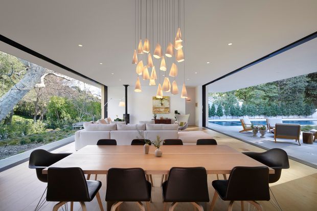Open-plan contemporary living and dining space with a large timber dining table in the foreground, surrounded by dark upholstered chairs. A pale sectional sofa, fireplace, and sculptural cluster pendant light occupy the living area beyond. Floor-to-ceiling glazed openings connect the room to a landscaped garden and pool terrace, creating a bright, spacious indoor-outdoor setting.