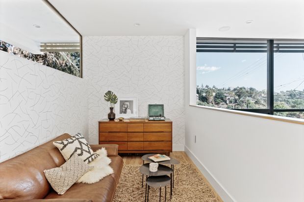 Bright mid-century-inspired living room with a brown leather sofa, patterned and sheepskin cushions, a low black nesting coffee table, a vintage-style wooden dresser, and a textured beige rug. White walls feature an abstract linear pattern, while large black-framed windows provide expansive views of a leafy suburban landscape. The room has a clean, airy arrangement with warm timber and leather accents.