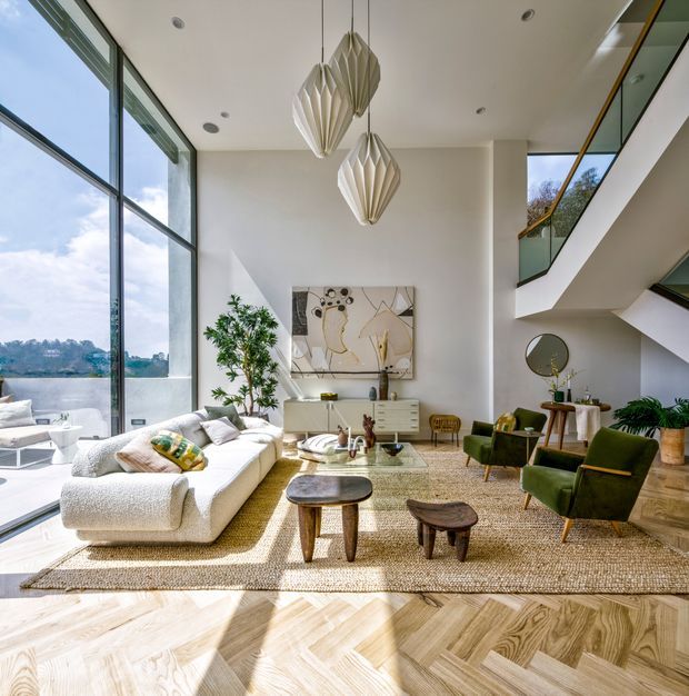 Bright, double-height living room with expansive floor-to-ceiling glazing, a white sectional sofa, green lounge chairs, timber coffee tables, a textured area rug, sculptural pendant lights, and a contemporary floating staircase with glass balustrades. The room opens visually to a leafy exterior and water or landscape beyond.