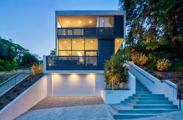 Contemporary multi-storey residence viewed from the street at dusk, with a projecting box-like form, extensive floor-to-ceiling glazing, dark horizontal privacy screens and balcony balustrades, recessed lighting, a wide garage, and a prominent external staircase leading to the upper levels. The home is integrated into a landscaped sloping site.