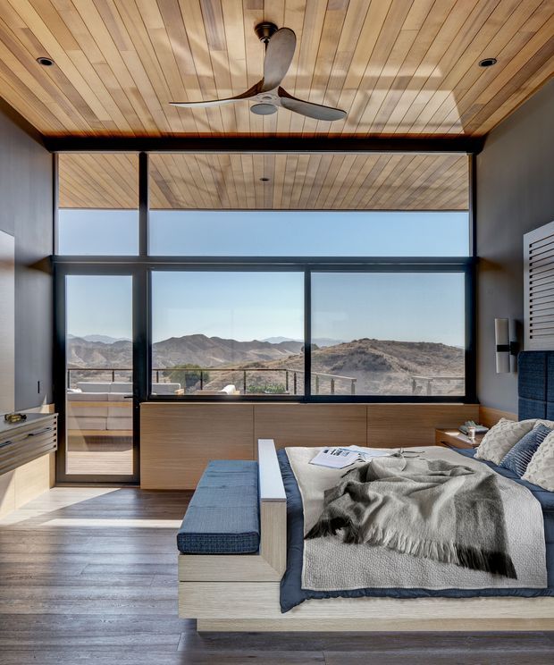 A contemporary bedroom with a built-in timber bed platform and upholstered blue bench, overlooking rugged mountainous terrain through expansive floor-to-ceiling glazing. The room features a warm timber-lined ceiling with an integrated ceiling fan, dark framed windows, timber flooring, built-in cabinetry, and a calm neutral-blue palette.