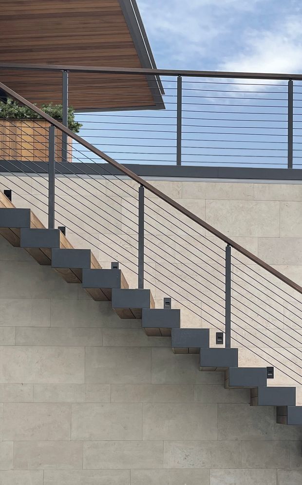 Contemporary exterior stairway with floating dark metal stair treads, slim horizontal cable balustrades, and a timber-lined overhanging roof or balcony above. Large pale stone or concrete wall panels form the backdrop, with a glimpse of sky and vegetation beyond.