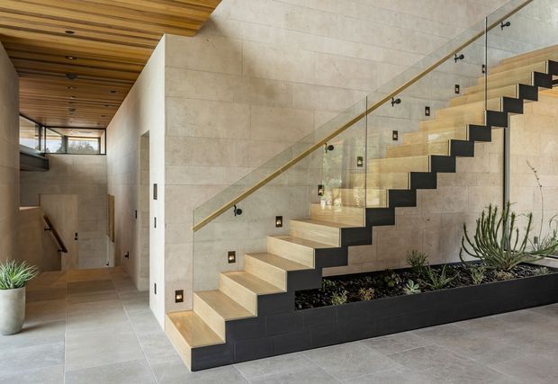 Contemporary interior stairway with a floating timber staircase, dark risers, glass balustrade, and timber handrail. Large-format beige stone or porcelain wall and floor tiles create a warm neutral backdrop, while a long built-in black planter with succulents runs beneath the stairs. A timber-lined ceiling, recessed lighting, and a partially visible lower-level hallway contribute to the refined modern design.