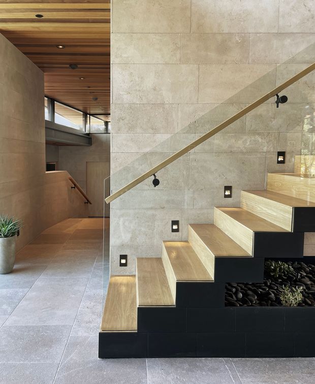 Interior architectural photograph of a contemporary stairway and landing. A floating timber-tread staircase with black built-in risers and planter niches rises beside a large pale stone-tiled wall, with a slim timber handrail and integrated stair lighting. The space continues into a circulation area with matching stone flooring and walls, a slatted timber ceiling, clerestory glazing, and a small indoor plant. The composition emphasises the staircase, material detailing, and warm natural light.