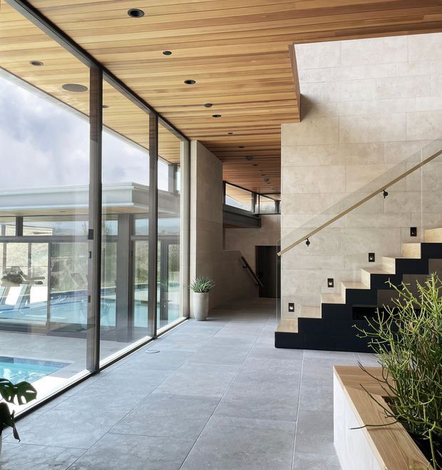 A refined contemporary interior hallway and stair zone with full-height black-framed glazing overlooking an indoor pool courtyard. The space features a warm timber-lined ceiling with recessed downlights, large-format pale stone or porcelain floor tiles, a pale stone feature stair wall, and a striking black staircase with a slim metal handrail. Built-in planter seating and sparse greenery add a natural accent to the open, light-filled space.