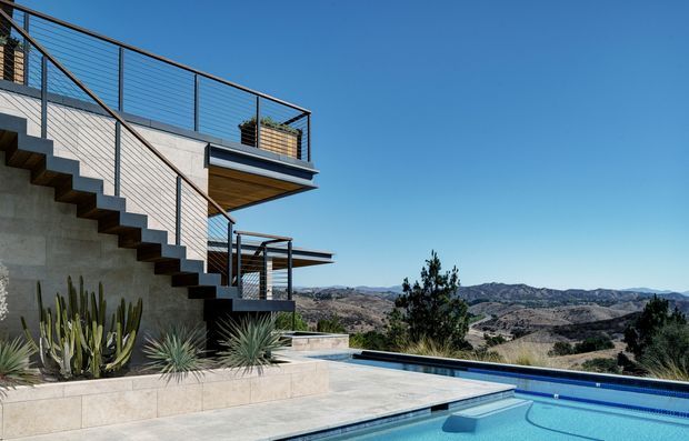 Modern multi-level hillside residence with exposed concrete walls, cantilevered balconies, open metal staircases and slender railings. A blue swimming pool and pale stone terrace occupy the foreground, while drought-tolerant landscaping and expansive mountainous terrain frame the view under a clear sky.