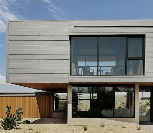Exterior view of a contemporary two-storey residence with a projecting upper volume clad in horizontal pale grey panels, extensive dark-framed glazing, and a warm timber soffit. The lower level combines large glazed openings with a timber-clad garage or outbuilding. Gravel landscaping with drought-tolerant planting surrounds the home, and a dry, rugged landscape is visible beyond.