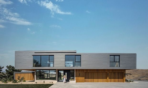 A contemporary two-storey residential house viewed from the front, with a long rectangular form, low-profile flat roof, horizontal grey cladding, extensive dark-framed glazing, warm timber-clad ground-floor sections, and an integrated double garage. A paved approach leads to the glazed entry, with sparse landscaping and a dry hillside setting.