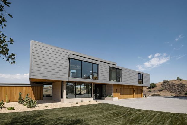 Modern two-storey residential house viewed from the front, with a strong horizontal composition, projecting upper volume, grey horizontal cladding, warm timber-lined lower facade, extensive dark-framed glazing, and a broad paved forecourt. The landscaped foreground includes lawn and sparse drought-tolerant planting, while a dry sloping site is visible to the side.