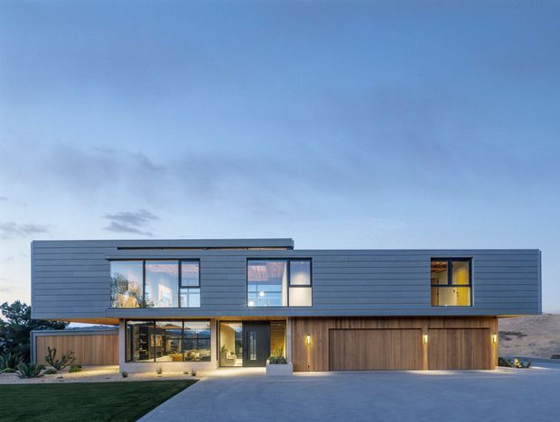 Contemporary two-storey residence viewed from the front at dusk, with a long rectangular upper volume clad in dark horizontal boards, extensive glazing, and a warm timber-clad lower level. The facade includes a sheltered entry, integrated garage doors, landscaped planting, and a broad concrete driveway beneath an open blue sky.