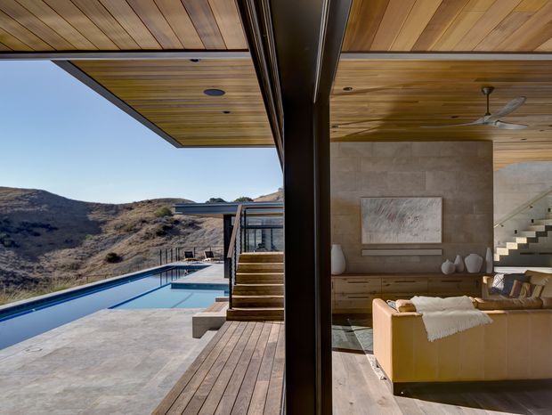 A contemporary indoor-outdoor living area opens directly onto a swimming pool and timber deck, with expansive glazing framing a rugged hillside landscape. The interior features a warm timber-lined ceiling, stone-clad feature wall, built-in timber cabinetry, a cream sofa, and a sculptural open staircase. A dark structural divider separates the living room from the pool terrace.