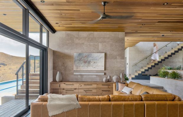 A warm, contemporary living room with a large tan leather sectional sofa facing a stone feature wall and low timber media console. Full-height glazing opens toward an exterior pool and rugged landscape, while a timber-lined ceiling with recessed lighting and a ceiling fan spans the room. A modern open stair with glass balustrade leads upward at the rear, with built-in planter joinery beneath it.