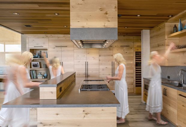 A warm, contemporary timber kitchen with extensive pale wood cabinetry, a large central island, integrated shelving, a cooktop beneath a substantial timber-and-metal rangehood, and a sink along the right wall. The ceiling is clad in matching timber boards with recessed downlights. Several blurred figures are present, but the kitchen joinery and finishes dominate the image.